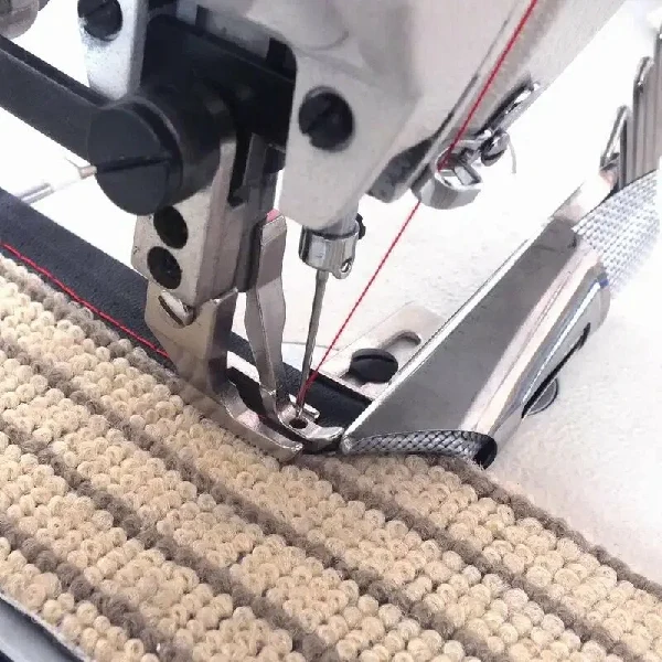 Industrial sewing machine applying black binding with red thread to a carpet edge.