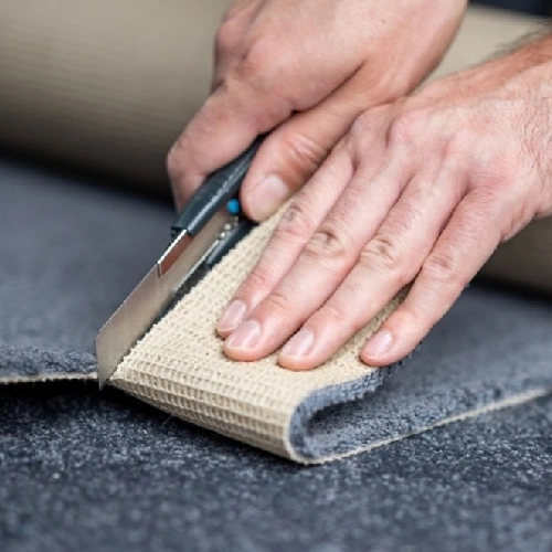 Using a utility knife to precision cut the backing of a gray carpet.