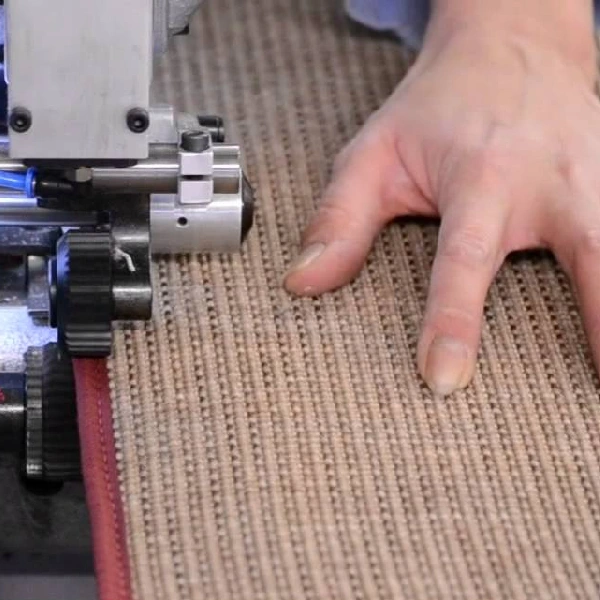 close-up of a carpet stitching machine stitching a red edge onto a tan rug.