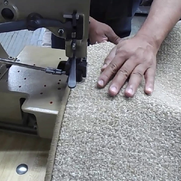 worker guiding a beige textured carpet through an industrial serging machine.