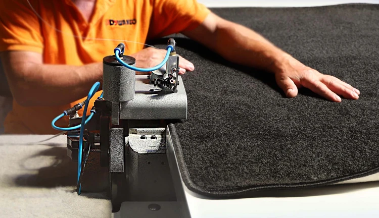 worker using an industrial serging machine to finish the edge of a black rug.